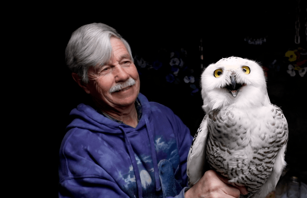 An older man with white hair and mustache wearing a blue hoodie smiles next to a white snowy owl with yellow eyes. The owl is perched on what appears to be the man's hand or arm, and both are shown against a black background.