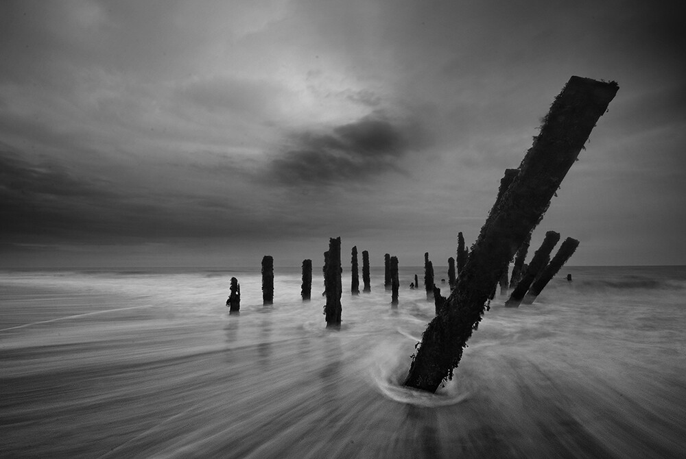 Atmospheric, long exposure, black and white photo of backwash moving around sea groynes under an overcast sky.