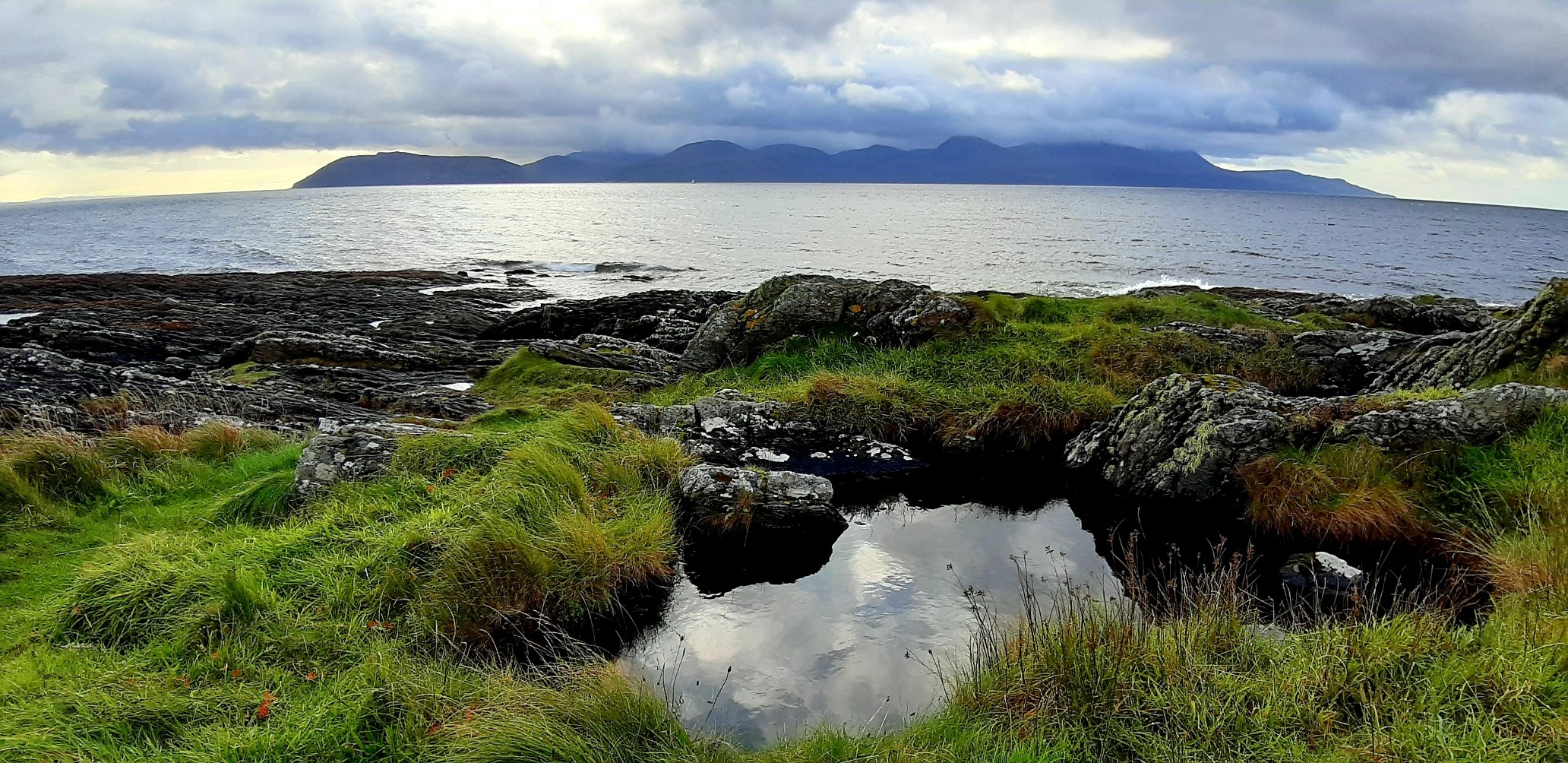 Looking across Kilbrannan Sound from Claonaig to the Isle of Arran. A pool in the foreground, surrounded by grass and schist, reflects the cloudy sky.