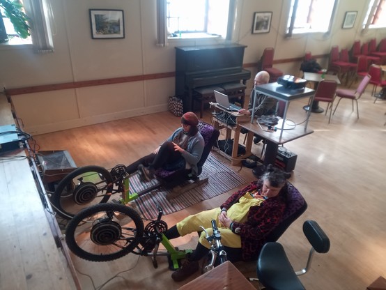Powering the pedal-powered cinema. Two people sit in (relatively) easy chairs, each with the cranks of  a bastardised bicycle in front of them. These bicycles have large hub motors which have been rewired to convert them into generators.