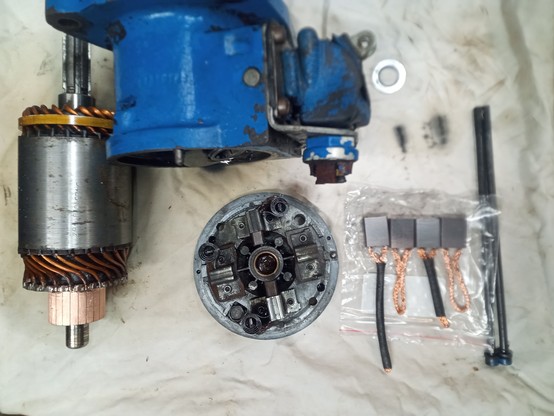 Parts of the starter motor laid out on a bench. The rotor is bottom left, the brush holder bottom centre, the set of four new brushes bottom right, next to the two long machine screws that will hold the whole assembly together.