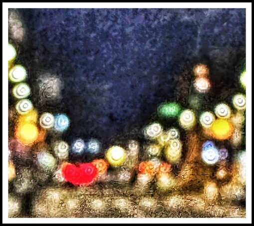 An out of focus photograph of a street. Dark, midnight blue sky with circles of light in a variety of bright colours - yellow, light blues, greens and whites. A red light in lower left of picture has blurred into a heart shape. 