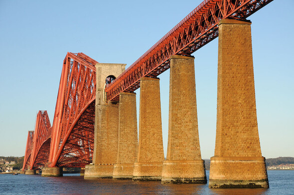 The Forth Bridge. The image shows the Forth Rail Bridge from the north-west in evening light, with the whole structure brightly lit, making the stonework look honey-coloured and the metalwork bright reddish-orange. The sky is blue.