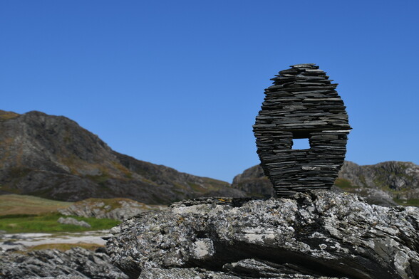 A stone sculpture, oblong shaped with a square window in the middle is standing on top of a large rocky outcrop on a beach. We see hills beyond and a blue sky. The sculpture is made from layers of thin slate like dark stones.