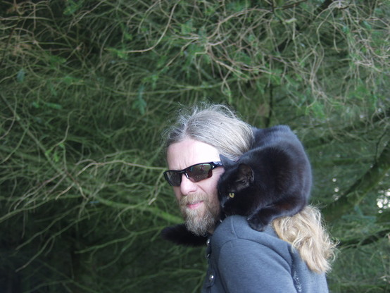 Penny sitting on my shoulder in a very apex-predator pose, looking intently at something to the left of the picture. I am wearing a grey hoodie and very racy cycling sunglasses. My hair is long. Behind us the trees are still thick and dark.