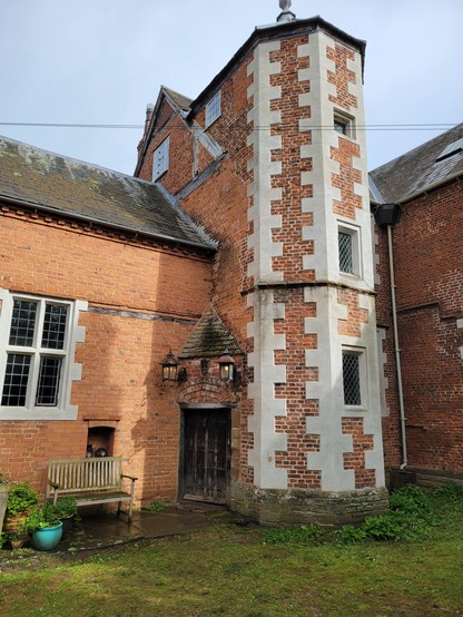 An external view of medieval Hellens Manor showing a brick tower with stone long-and-short quoins and the outside of the hall. There is a super-cute little doorway across the corner where the hall and tower meet, with a tiled sloped roof, lanterns and a brick arch over it. The door is wooden and very old.