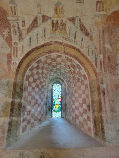 A stunning medieval wall painting around a Norman window. The window is recessed with a splayed frame and ledge. The underside of the arch is decorated with a red and white chequerboard pattern, and above the arch there is a scene of rooftops. The window itself is a slim arched depiction of (probably) St George.