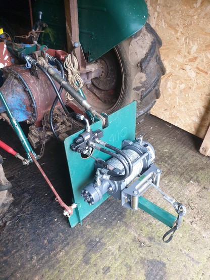 Winch on the tractor, view from the left rear quarter. The heavy winch with its steel cable are mounted to the plate. It is linked by two hydraulic hoses to a control valve mounted on the top left of the plate. The plate is mounted by the frame to the three point linkage on the tractor, and the valve is linked by two more hydraulic hoses to the hydraulic circuit of the tractor.