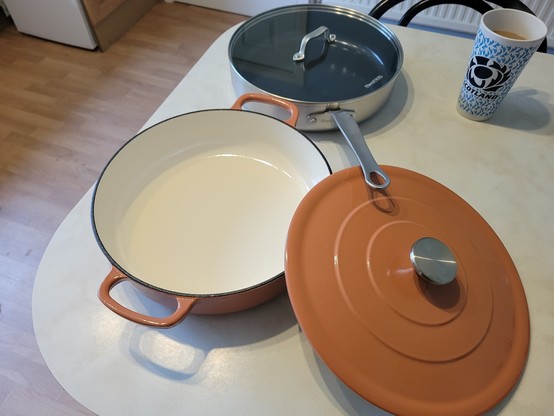 Two saucepans, at the front a Le Creuset-style cast iron and enamel two-handled casserole dish with its lid off showing you its beautiful cream enamel interior, at the back a stainless steel heavy based frying-type pan with a long handle and clear lid.