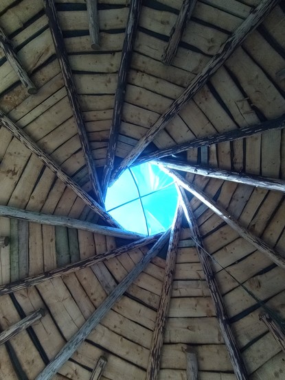 The view up. Through the circular eye in the crown of the roof, a clear blue sky is visible. Around it, the rough wooden rafters -- debarked tree trunks -- radiate tangentially, while the sarking boards spiral in the opposite direction.