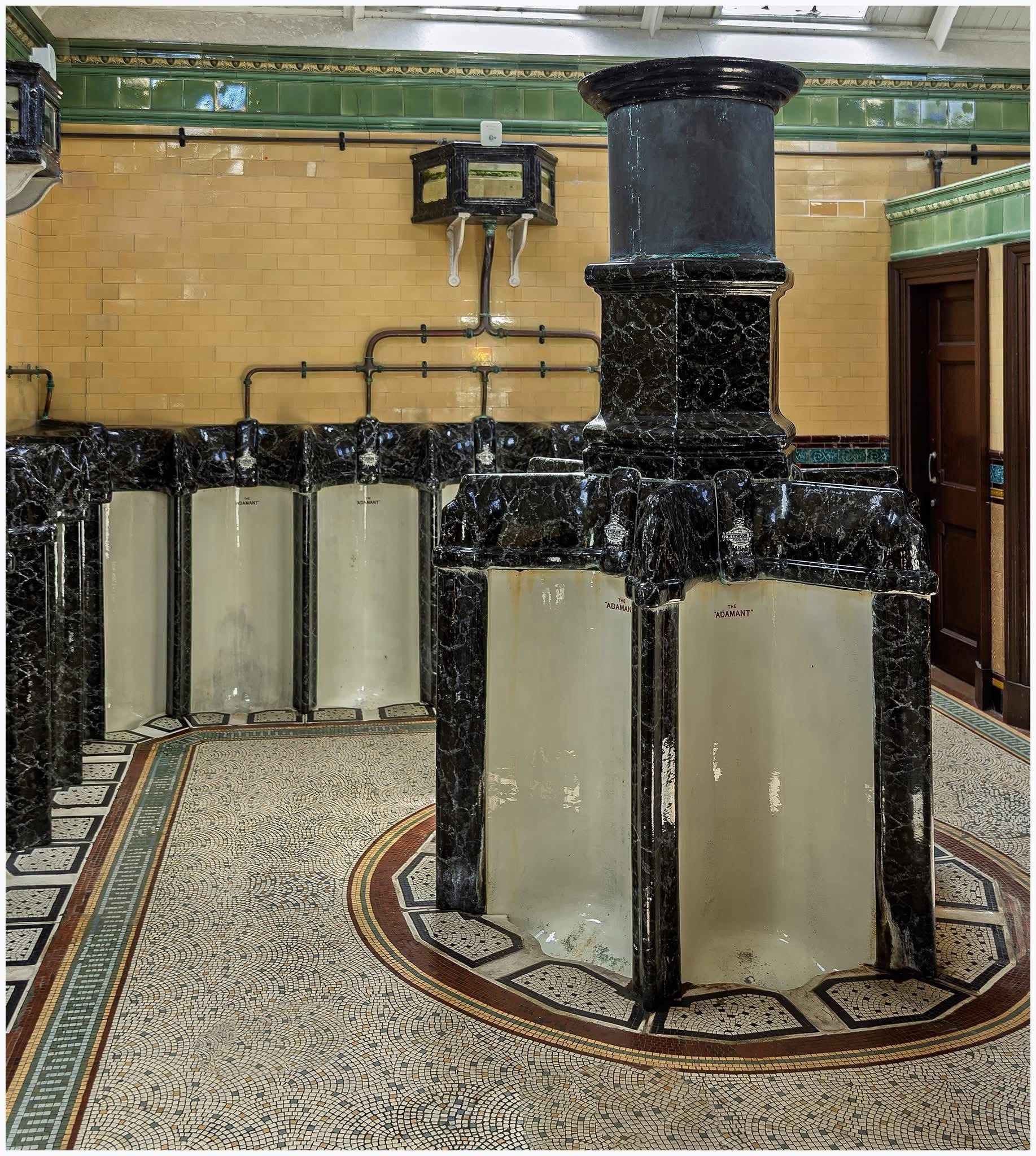 Historic restroom featuring ornate black marble fixtures and a tiled floor with intricate patterns. The central structure is prominent, surrounded by several partitions. The walls are decorated with yellow tiles and green accents. Historic restroom featuring ornate black marble fixtures and a tiled floor with intricate patterns. The central structure is prominent, surrounded by several partitions. The walls are decorated with yellow tiles and green accents.