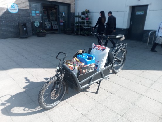 The big grey bakfiets-style bike, loaded with a white sack of cattle sweeties, a blue container of petrol, and a lot of groceries, in bright sunlight outside the co-op in Castle Douglas.
