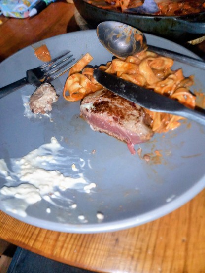 A grey plate on a wooden table. On the plate, a piece of fine beef, seared on the outside and almost raw in the middle. Behind the beef, linguine in an orange coloured sauce. In the foreground, a smear of white horseradish sauce.