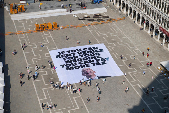 Aerial photo of the Piazza San Marco in Venice, showing a very large unfurled banner that reads "If you can rent Venice for your wedding, you can pay more tax" with a photo of the owner of Amazon.