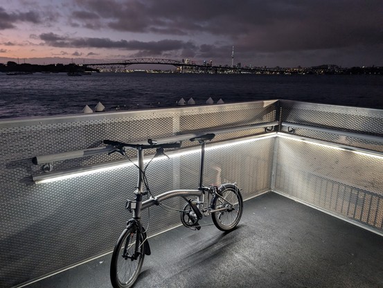 Brompton bike against the railings of an urban ferry terminal with Auckland city skyline in the background at dawn 
