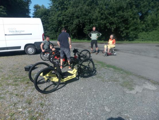 Two hand-cycles, two people sitting in wheelchairs, and various other people standing around in a parking area.