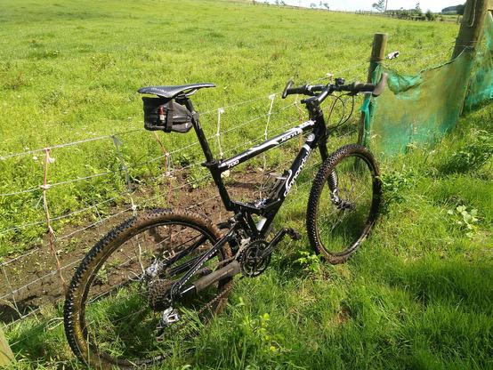 My 2004 Cannondale Jekyll, a lightweight black full suspension mountain bike with a 'lefty' single sided front fork, leaning against a fence in a green field.