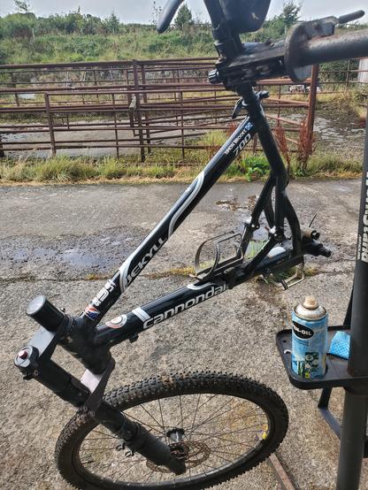 The same bike, hanging from my workstand, with its rear wheel, rear triangle, and handlebars removed. The workstand stands in a concreted roadway, with the rusty railings of the old communal cattle management yard behind.