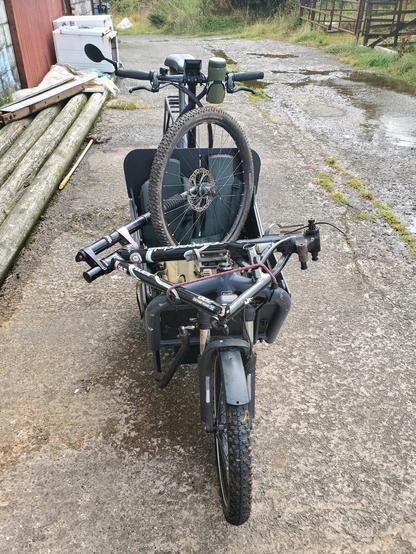 Transporting what's left of the bike down to the bike workshop in the village, on another bike -- the big black cargo bike -- of course. The cargo bike faces the camera, standing on the concrete roadway. The frame and front wheel of the mountain bike lie across its box.