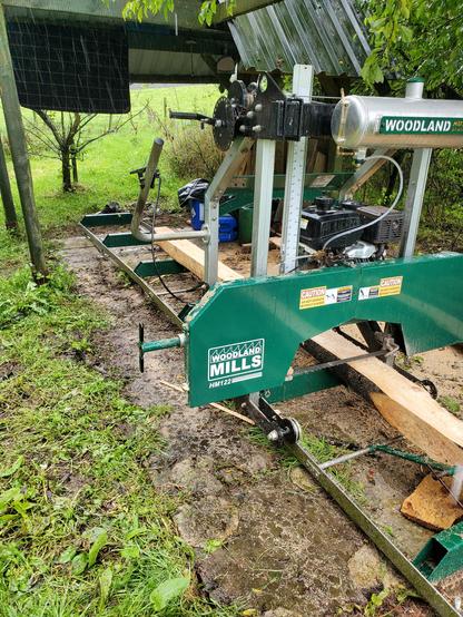 My sawmill, cutting a not very big, not very straight log on a very wet day. The log lies on the bed of the mill, mostly squared up. The carriage of the sawmill, with its green steel box enclosing the blade, sits across it.