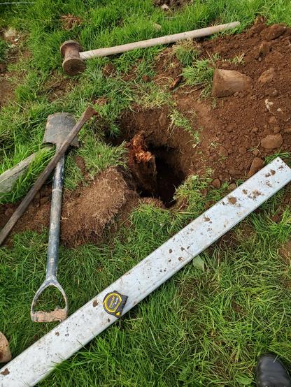 The remains of a rotten, broken gate post stand in the ground. Around it a hole half a metre deep has been dug, and the tools used to dig it are scattered on the grass.

In front of the hole, a new, galvanised steel post lies waiting to go in — but the hole is not deep enough yet.